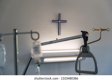 Religious Blue Cross On Focus In A White Hospital Room. Medical Equipment With A Religious Symbol Hanging On The Wall And Lit From Below. Empty Hospital Room With A Iv Pole, Handle And Fluorescent.