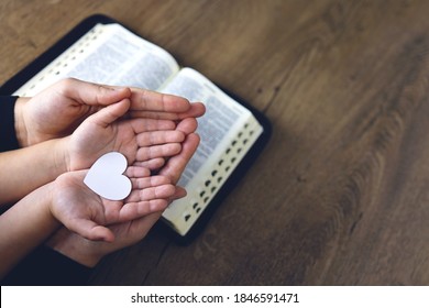 Religious Christian girl praying with her mother indoors. Bible in background. Hads holding white paper heart. Space for text
