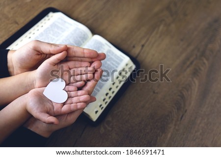 Religious Christian girl praying with her mother indoors. Bible in background. Hands holding white paper heart. Space for text
