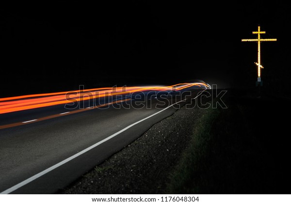 Religious Cross on the Side of Road in the\
light of the headlights of passing cars.\
Night.
