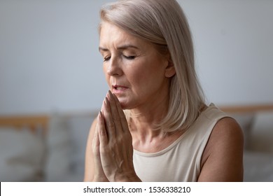 Religious faithful middle aged woman praying with hope faith holding hands clasped together in namaste at home in morning, old mature lady believer christian saying worship prayer with eyes closed
