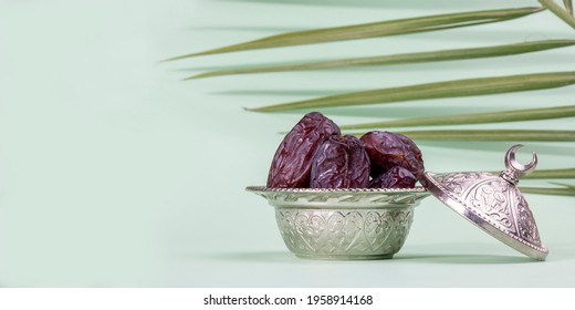 Religious Islamic festival and holy month of Ramadan, EID KAREEM, concept: A silver metallic and ornate bowl with dried date fruits. Palm leaves and green background with copy space 