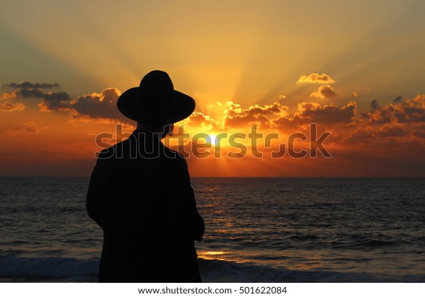 Religious Jew\
standing on the coast of Mediterranean sea in Israel and watching\
the sunset, deep in\
thought.