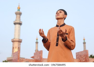 Religious middle eastern man in brown muslim shirt and black cap standing against mosque background, praying earnestly with his hands raised and looking up