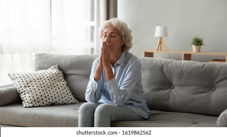 Religious Older Woman Praying With Hope, Holding Hands Together In Prayer, Sitting On Couch At Home Alone, Thoughtful Mature Female With Closed Eyes Meditating, Feeling Gratitude, Making Wish