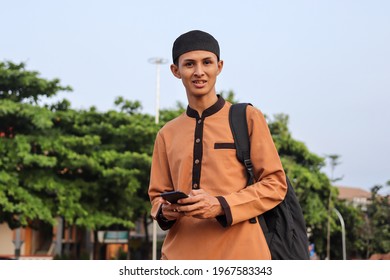 Religious smiling middle eastern man in brown muslim shirt and black cap carrying a bag, using his mobile phone, and standing against mosque background