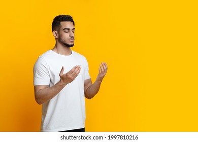Religious young arab muslim man with closed eyes praying, holding palms face up, whispering pray, yellow studio background, panorama with copy space. Religion, islam, believing concept