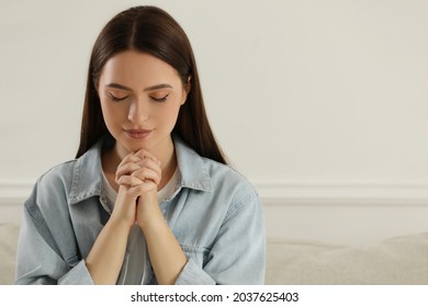 Religious young woman with clasped hands praying indoors