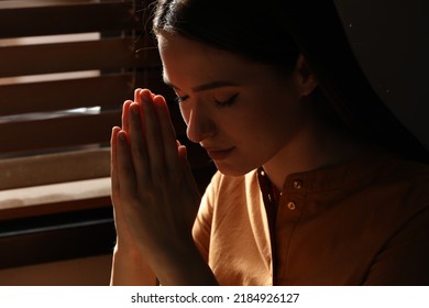 Religious young woman with clasped hands praying indoors, closeup