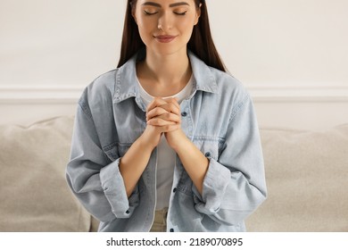 Religious young woman with clasped hands praying indoors, closeup