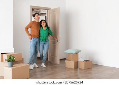 Relocation Concept. Happy Millennial Newlyweds Walking In New Empty House With Cardboard Boxes On Floor, Young Family Of Two People Moving Into Bought Apartment, Excited Guy And Lady Choosing Flat