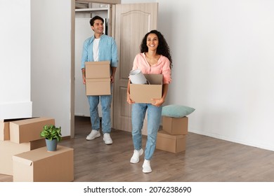 Relocation Concept. Happy Millennial Newlyweds Holding Cardboard Boxes Walking In New House, Young Family Of Two People Moving Into Bought Apartment, Excited Guy And Lady Carry Personal Belongings