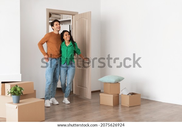 Relocation Concept. Happy millennial newlyweds\
walking in new empty house with cardboard boxes on floor, young\
family of two people moving into bought apartment, excited guy and\
lady choosing flat