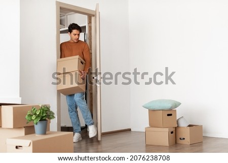 Relocation Concept. Portrait of young millennial guy holding pile of cardboard boxes walking in new house, busy person with full hands moving inside bought apartment, opens door with leg