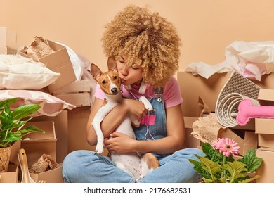 Relocation And Ownership Concept. Curly Haired Young Woman Moves To New House Or Apartment Embraces And Kisses Pet With Love Sits Crossed Surrounded By Carton Boxes Full Of Personal Belongings