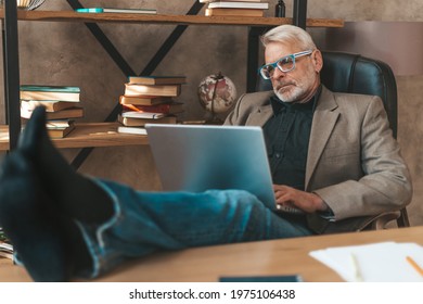 Reluctance To Work. Senior Man Sits With Laptop, Legs Propped Up On Desk In Office. Procrastination.