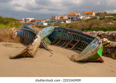 remains of blue and green boats sunk in the sand on the beach in the background of the river forest mass and fishing village cloudy sky - Shutterstock ID 2251545909