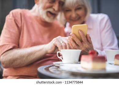 Remember we. Focus on male hands holding smartphone and showing it to old woman. They are sitting with delight and looking at screen while drinking tea outside - Powered by Shutterstock