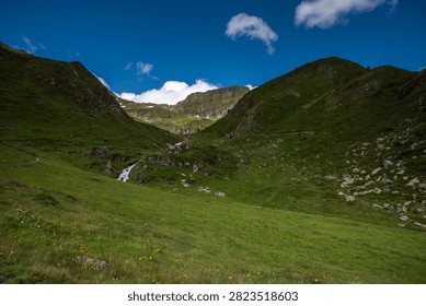 A Remote Alpine Valley Opens Between Green Mountain Slopes Beneath a Deep Blue Sky - A broad alpine valley stretches between steep grassy mountainsides covered with low vegetation, exposed rock, and scattered wildflowers. A small stream cascade is visible in the middle distance, descending through the terrain toward the valley floor. Patches of lingering snow remain tucked beneath higher ridges, indicating the elevation of the landscape despite summer conditions. Bright sunlight enhances the vivid greens of the grassland and contrasts with the intense blue sky overhead. The image captures the openness, solitude, and natural beauty of a remote mountain environment.