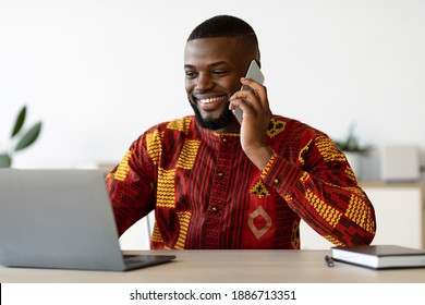 Remote Career. Young African Freelancer Man In Traditional Clothes Talking On Cellphone And Working On Laptop Computer At Home Office, Having Pleasant Conversation, Enjoying Distance Job, Copy Space