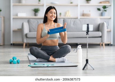 Remote Coaching Concept. Smiling Asian Female Fitness Trainer Recording Video Tutorials At Home, Fit Korean Sport Blogger Showing Exercises With Resistance Band At Camera, Using Smartphone On Tripod - Powered by Shutterstock