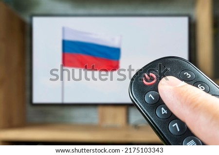 Remote control in a man's hand on the background of a TV and a Russian flag. The concept of disconnecting Russian channels in Europe and the USA. Blocking of Russian TV broadcasting.