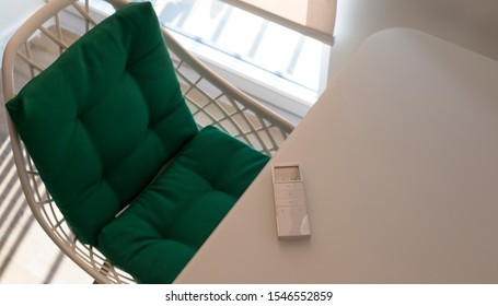 Remote Control Panel For Automatic Roller Blinds Lies On A Beige Table In The Living Room. Nearby Is A Chair With A Green Pillow. On The Window Roller Shades With Automatic Control.  