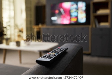 Remote control resting on sofa armrest with blurred television screen in background showing colorful content, modern living room setting with coffee table and shelves visible