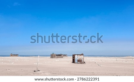 A remote desert area featuring scattered small structures and a rusted container on a sandy expanse, set against a vast blue sky, suggesting an isolated outpost