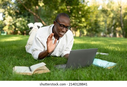 Remote Education Concept. Black Male Student Video Calling Via Laptop, Having Online Lecture And Waving Hello To Webcamera, Lying On Lawn In Park Or College Campus Outdoors