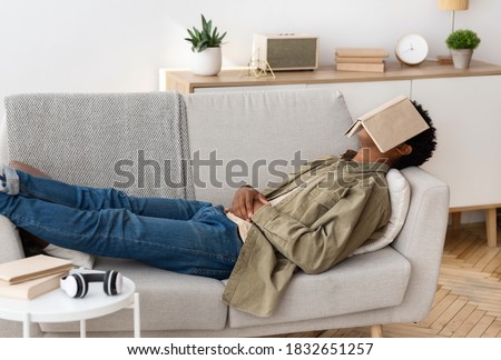 Remote education concept. Bored black teen student with book on his face sleeping on couch at home. African American teenager unwilling to do dull home assignment, napping on sofa with textbook