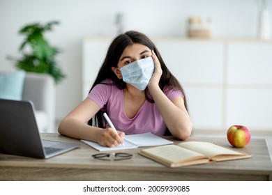 Remote Education During Coronavirus Concept. Indian Teen Girl In Face Mask Sitting At Table With Laptop, Taking Notes, Making Study Project For Online Course, Participating In Web Conference At Home