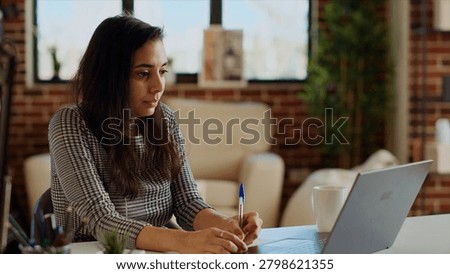 Remote employee at desk doing multitasking, transcribing data from laptop to notepad, Indian woman in apartment trying to complete job tasks before deadline, writing info on paper using pen.