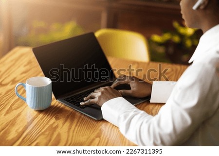 Remote job, freelance concept. Unrecognizable black woman in smart casual freelancer typing on laptop with black empty screen, using earpods, businesswoman working at cafe, high angle shot, mockup