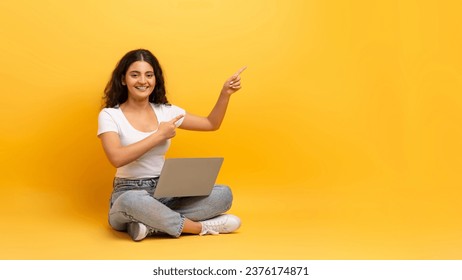 Remote job opportunities, freelance. Happy beautiful curly young indian woman student wearing casual outfit sitting on floor with laptop on her lap, pointing at copy space, yellow background, panorama