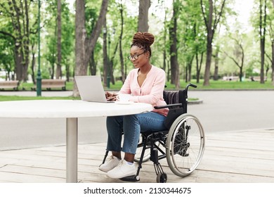 Remote Job For People With Disability. Cheerful Young Black Woman In Wheelchair Working Online, Using Laptop At Outdoor Cafe. African American Lady Studying Or Communicating On Web