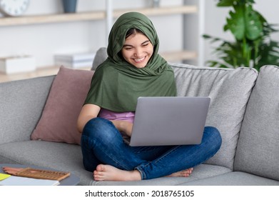 Remote learning concept. Happy Muslim teen girl in hijab studying online, sitting on sofa with laptop at home. Indian teenager in Islamic headscarf using PC, doing assignment, communicating to teacher