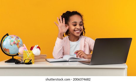 Remote Learning Concept. Portrait Of Positive African American Schoolgirl Sitting At Desk, Making Video Call Using Laptop Computer. Smiling Young Black Girl Waving Hand While Having Virtual Class