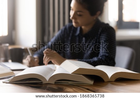 Remote learning. Diligent millennial mixed race female student prepare for exam at home. Confident indian woman write up notes thesis for research report essay. Focus on opened paper books on table