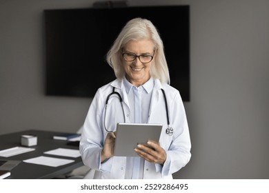 Remote medical care. Smiling pleasant older adult woman doctor work on tablet pc online support patient on distance. Senior lady healthcare expert doc use pad in telemedicine give advice by video call - Powered by Shutterstock