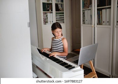 Remote Music Lesson, A Child Girl Playing The Digital Piano And Looking At A Laptop, Online Learning And Video Chat On Learning To Play The Piano. Authentic Lifestyle In A Real Interior