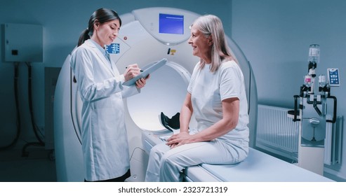 Remote shooting. Female doctor is standing near patient and explaining procedure. Female patient is smiling and listening to doctor carefully. Medical worker is holding tablet and talk to patient.