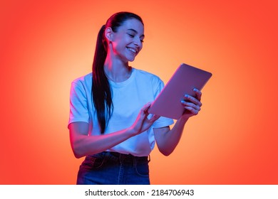 Remote studying. Portrait of young emotional girl, student in white t-shirt isolated on orange color background in neon light. Concept of beauty, art, fashion, education - Shutterstock ID 2184706943