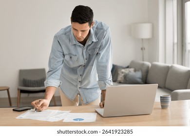 Remote work and data analysis. Busy gen z male financial analyst accountant freelancer stand by home office desk hold pen focused on reviewing paper documents prepare statistic report using notebook