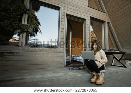 Remote work and escaping to nature concept. Woman works on laptop against tiny cabin house.