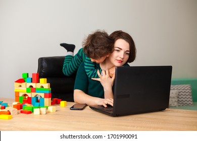 remote work at home. the son, the child sits out of boredom at the tired mother's neck, demanding the attention of the parent. A woman works at home at a computer