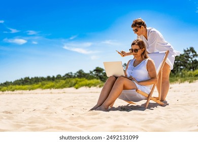 Remote work on beach. Two young women dressed in white: one sitting on deckchair and holding laptop and the other standing behind her with smartphone in her hand on sandy beach on sunny day. - Powered by Shutterstock