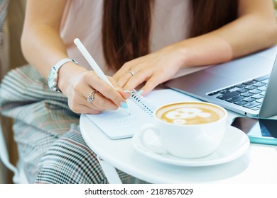 Remote Work Outside The Office, Outdoor Workplace. Desktop With A Cup Of Cappuccino, Laptop And Female Hands Holding Pen And Writing In A Notebook.