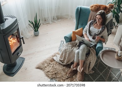 Remote Work. Young Woman Using Laptop In A Cozy Armchair And Plaid Sitting By The Fireplace With A Domestic Cat And Drinking Tea At Home