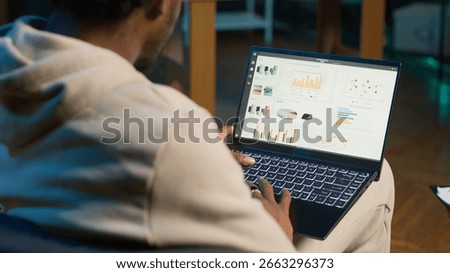 Remote worker accountant doing math calculations using financial graphs on laptop, working at home. Self employed guy in loft comparing business economic figures on notebook. Camera A.